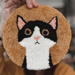 Può includere: Un tappeto rotondo fatto a mano con la faccia di un gatto. Il gatto è bianco e nero con orecchie e naso rosa e occhi neri. Lo sfondo è beige. Il tappeto è realizzato in un materiale strutturato.