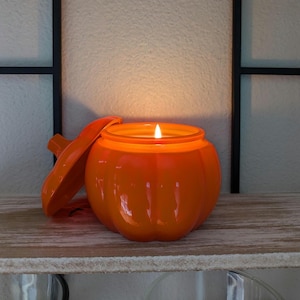 May include: An orange pumpkin-shaped candle with a lit wick and a matching lid. The candle sits on a wooden shelf, with a black metal frame in the background. The candle is a vibrant orange color, perfect for fall decor.