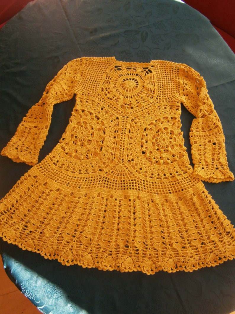 May include: A mustard yellow crocheted dress with a round, lacy design in the center and long sleeves.