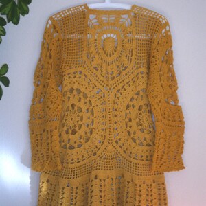 May include: A mustard yellow crochet cardigan with a long, flowing silhouette and intricate lace detailing. The cardigan has a wide, open neckline and long sleeves.