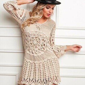 May include: A beige crochet dress with a scalloped hem and long sleeves. The dress has a lace-like pattern and a tie at the neckline. The model is wearing a black fedora hat.