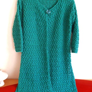 Puede incluir: Un cárdigan de ganchillo verde turquesa, hasta la rodilla, con cuello en V y mangas tres cuartos. El cárdigan presenta un patrón texturizado en forma de onda y un cierre de un solo botón azul redondo. La prenda está colgada de una percha negra sobre un fondo blanco.