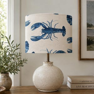 May include: A table lamp with a cream-coloured, textured, spherical ceramic base and a cylindrical lampshade featuring a repeating pattern of blue lobsters on a light beige background. The lamp has a simple metal neck connecting the base to the shade.