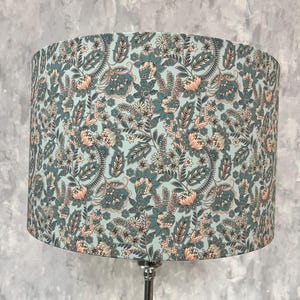 Pode incluir: Abajur cil&iacute;ndrico com padr&atilde;o floral. O abajur apresenta um fundo azul claro com um design repetido de flores, folhas e formas de paisley em tons de verde-azulado, p&ecirc;ssego e creme. Montado em uma base de metal prateado.