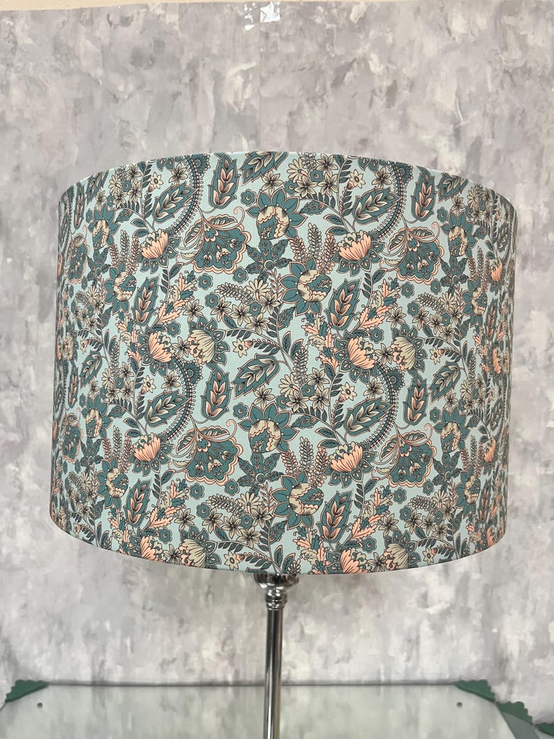 Pode incluir: Abajur cil&iacute;ndrico com padr&atilde;o floral. O abajur apresenta um fundo azul claro com um design repetido de flores, folhas e formas de paisley em tons de verde-azulado, p&ecirc;ssego e creme. Montado em uma base de metal prateado.