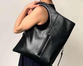 Puede incluir: Un bolso tote de cuero negro con un asa de corte circular único. El bolso tiene una correa larga y un bolsillo con cremallera en la parte delantera.