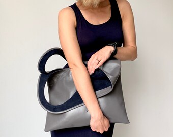 May include: A gray leather tote bag with a unique design. The bag has a large, open top and a flap that can be folded over to close it. The bag is held by a person wearing a black tank top.