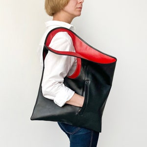 Black leather shoulder bag with red interior collar is finished with an outside vertical zipper pocket. The bag is fully lined and has two inner pockets. The bag can be carried on the shoulder or as a clutch. Height 19 Width 17 inches.