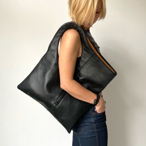 Black leather shoulder bag with tan interior collar. It has a rectangular shape with an oval armhole holder. The bag has an outside vertical zipped pocket. The bag can be carried in hand as a tote, over the shoulder or as a fold over clutch.