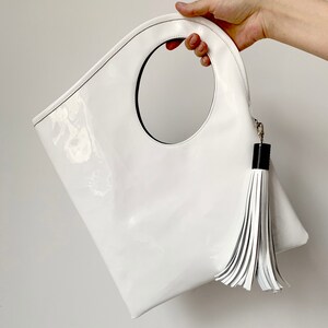 White patent leather bag is finished with an outside vertical zipper pocket and a strap. The bag is fully lined and has two inner pockets. The bag can be carried on the shoulder or crossbody. Height 15 Width 13.5 inches.