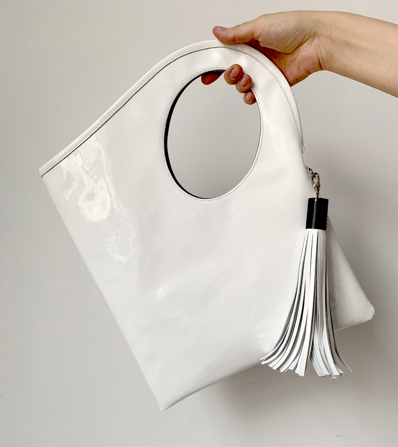 White patent leather bag is finished with an outside vertical zipper pocket and a strap. The bag is fully lined and has two inner pockets. The bag can be carried on the shoulder or crossbody. Height 15 Width 13.5 inches.