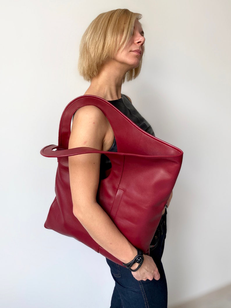 Burgundy leather bag has a rectangular shape. It is finished with an outside vertical zipper pocket. Height 17.5, Width 15.5 inches. The bag is fully lined and has two interior pockets. It can be carried on a shoulder or as a folded clutch.
