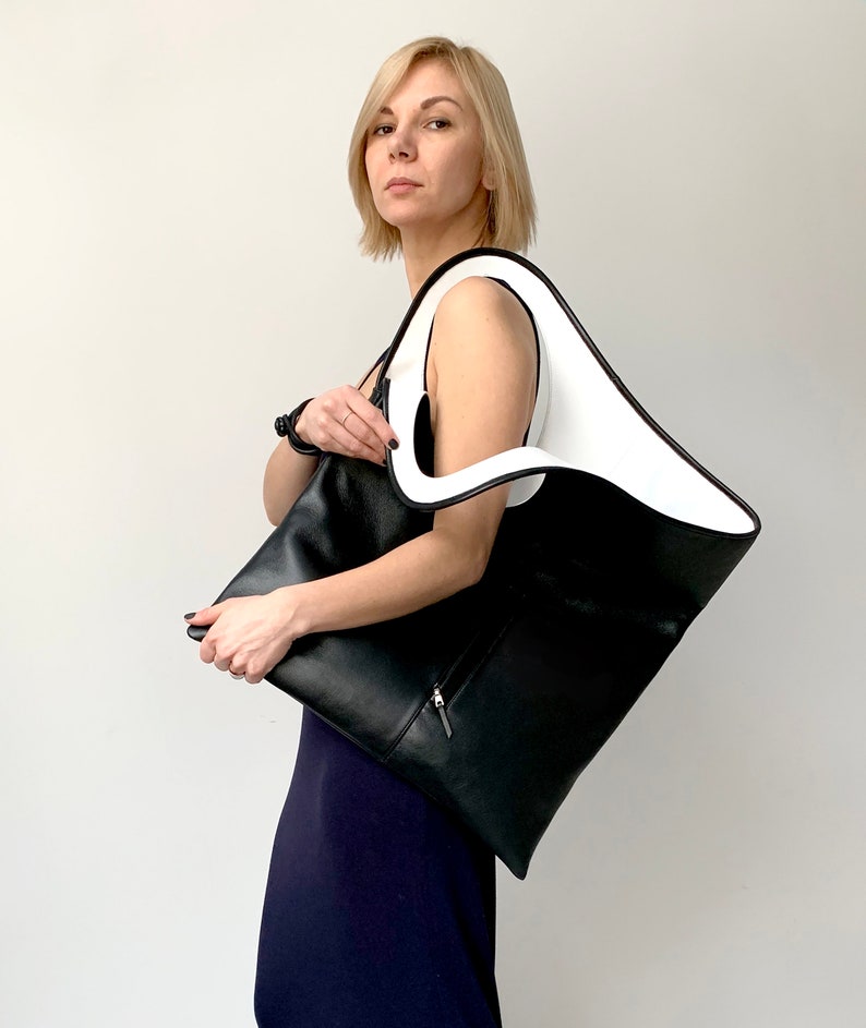 Black leather bag is finished with an outside vertical zipper pocket and a strap. The bag is fully lined and has two inner pockets. The bag can be carried on the shoulder or crossbody. Height 20.5 Width 18.5 inches.