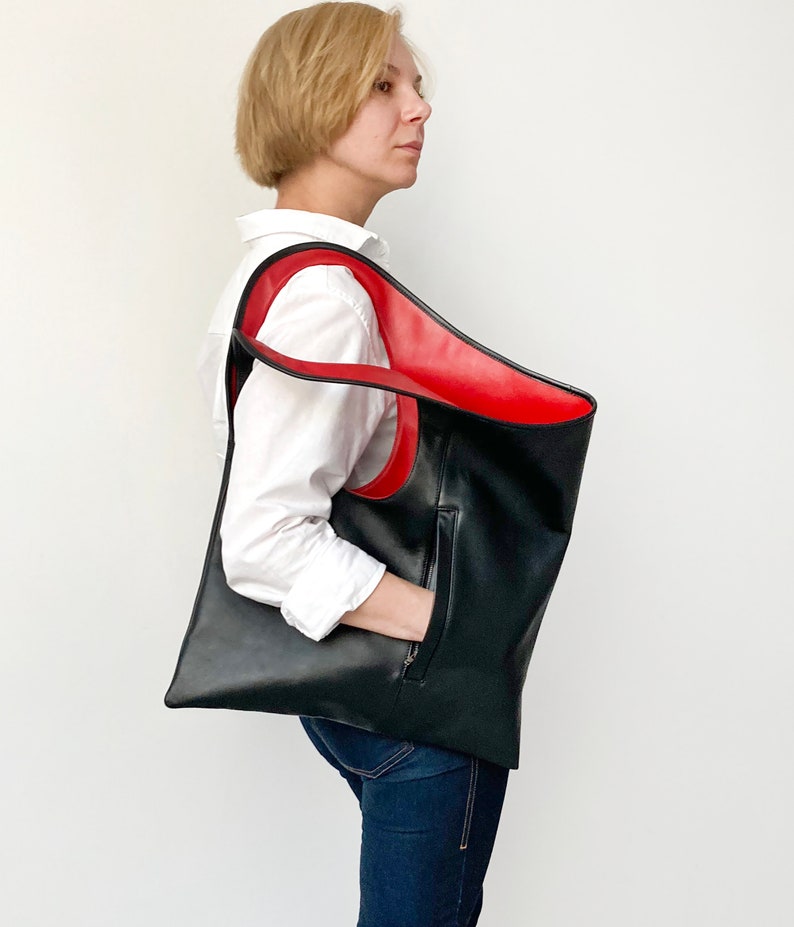 Black leather shoulder bag with red interior collar is finished with an outside vertical zipper pocket. The bag is fully lined and has two inner pockets. The bag can be carried on the shoulder or as a clutch. Height 19 Width 17 inches.