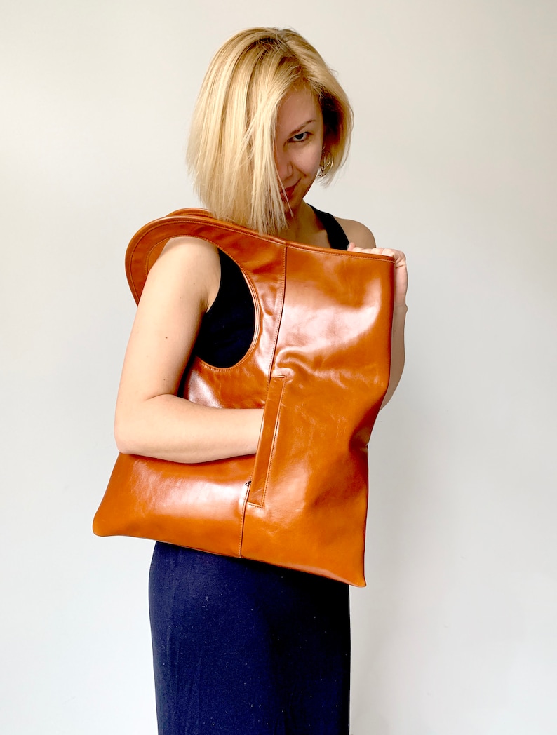 Cognac leather bag is finished with an outside vertical zipper pocket and a strap. The bag is fully lined and has two inner pockets. The bag can be carried on the shoulder or crossbody. Height 17.5 Width 15.5 inches.