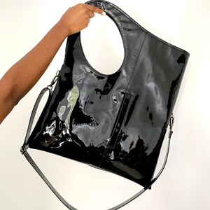 Black patent leather shoulder bag is finished with an outside vertical zipper pocket and an adjustable strap. The bag is fully lined and has two inner pockets. The bag can be carried on the shoulder or crossbody. Height 19 Width 17 inches.