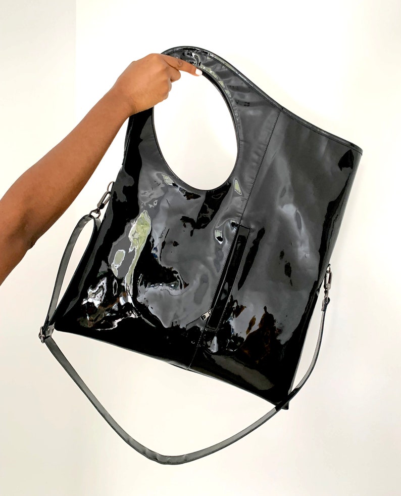 Black patent leather shoulder bag is finished with an outside vertical zipper pocket and an adjustable strap. The bag is fully lined and has two inner pockets. The bag can be carried on the shoulder or crossbody. Height 19 Width 17 inches.