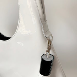 White patent leather bag is finished with an outside vertical zipper pocket and a strap. The bag is fully lined and has two inner pockets. The bag can be carried on the shoulder or crossbody. Height 15 Width 13.5 inches.