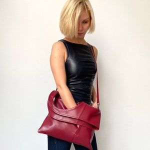 Burgundy leather bag has a rectangular shape. It is finished with an outside vertical zipper pocket and a strap. Height 17.5, Width 15.5 inches. It is fully lined and has two interior pockets. It can be carried on a shoulder or as a folded clutch.