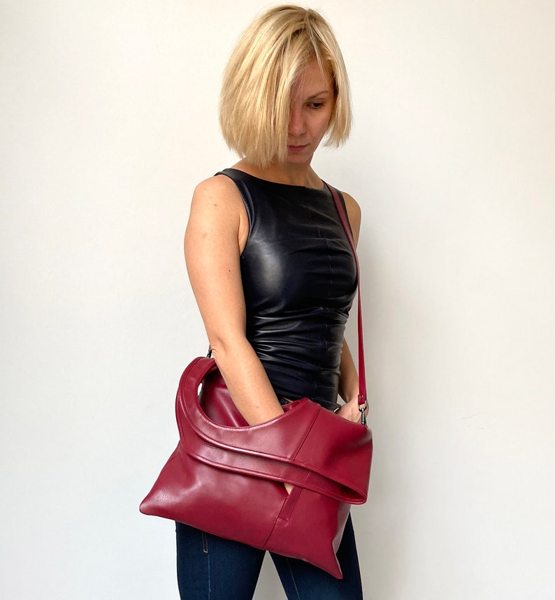 Burgundy leather bag has a rectangular shape. It is finished with an outside vertical zipper pocket and a strap. Height 17.5, Width 15.5 inches. It is fully lined and has two interior pockets. It can be carried on a shoulder or as a folded clutch.