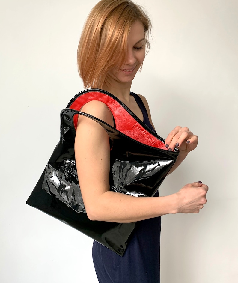 Black patent leather bag is finished with an outside vertical zipper pocket and a strap. The bag is fully lined and has two inner pockets. The bag can be carried on the shoulder or crossbody. Height 15 Width 13.5 inches.