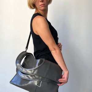 Grey leather bag has a rectangular shape. It is finished with an outside vertical zipper pocket and a strap. Height 19, Width 17 inches. It is fully lined and has two interior pockets. It can be carried on a shoulder or as a folded clutch.