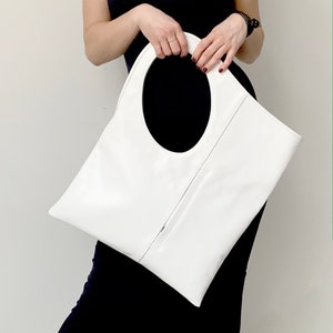 White leather bag is finished with an outside vertical zipper pocket and a strap. The bag is fully lined and has two inner pockets. The bag can be carried on the shoulder or crossbody. Height 17.5 Width 15.5 inches.