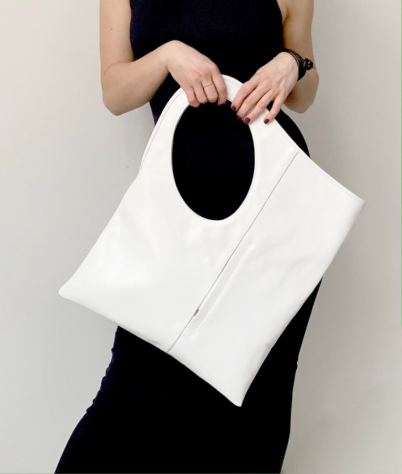 White leather bag is finished with an outside vertical zipper pocket and a strap. The bag is fully lined and has two inner pockets. The bag can be carried on the shoulder or crossbody. Height 17.5 Width 15.5 inches.