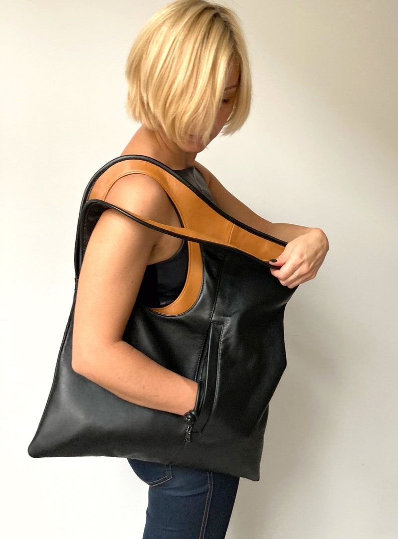 Black and tan leather bag has a rectangular shape. It is finished with an outside vertical zipper pocket and a strap. Height 19, Width 17 inches. It is fully lined and has two interior pockets. It can be carried on a shoulder or as a folded clutch.