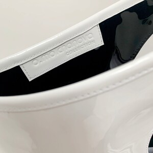 White patent leather bag is finished with an outside vertical zipper pocket and a strap. The bag is fully lined and has two inner pockets. The bag can be carried on the shoulder or crossbody. Height 15 Width 13.5 inches.