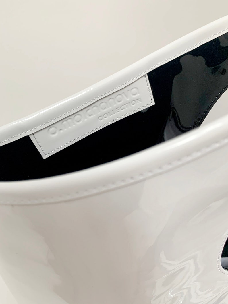 White patent leather bag is finished with an outside vertical zipper pocket and a strap. The bag is fully lined and has two inner pockets. The bag can be carried on the shoulder or crossbody. Height 15 Width 13.5 inches.