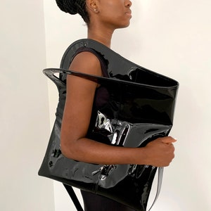 Black patent leather shoulder bag is finished with an outside vertical zipper pocket and an adjustable strap. The bag is fully lined and has two inner pockets. The bag can be carried on the shoulder or crossbody. Height 19 Width 17 inches.