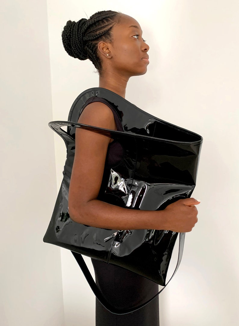 Black patent leather shoulder bag is finished with an outside vertical zipper pocket and an adjustable strap. The bag is fully lined and has two inner pockets. The bag can be carried on the shoulder or crossbody. Height 19 Width 17 inches.