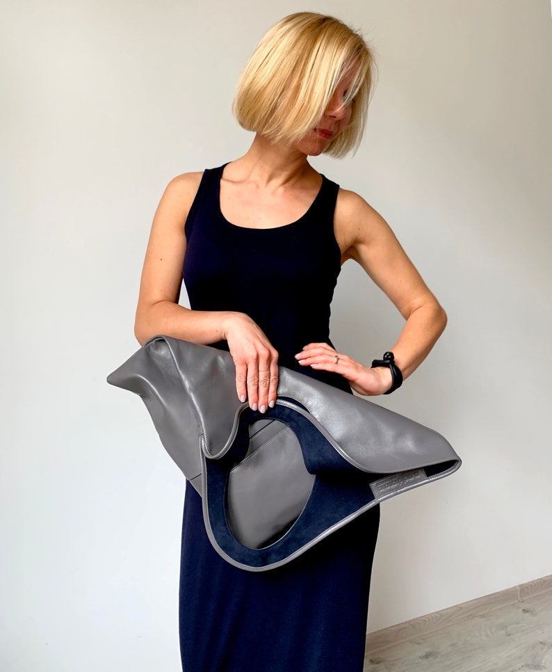 Grey leather bag is finished with an outside vertical zipper pocket and a strap. The bag is fully lined and has two inner pockets. The bag can be carried on the shoulder or crossbody. Height 17.5 Width 15.5 inches.
