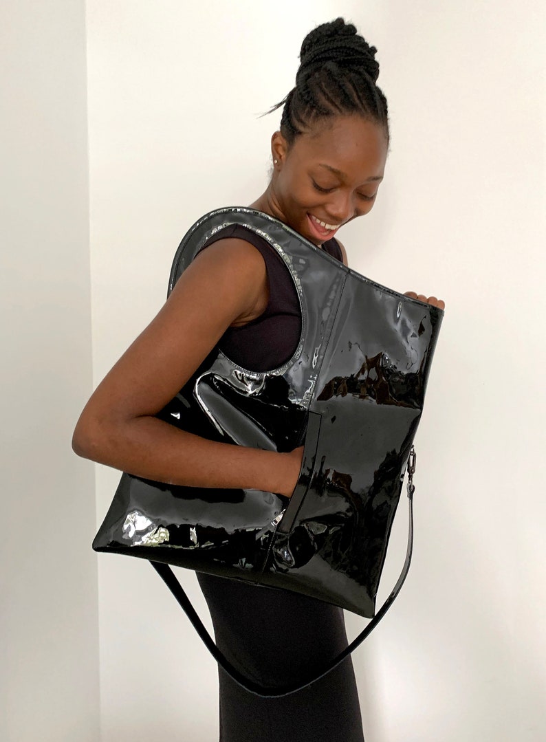 Black patent leather shoulder bag is finished with an outside vertical zipper pocket and an adjustable strap. The bag is fully lined and has two inner pockets. The bag can be carried on the shoulder or crossbody. Height 19 Width 17 inches.