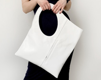 May include: A white leather tote bag with a large oval handle. The bag has a rectangular shape with a vertical zipper detail. The bag is held against a dark background.