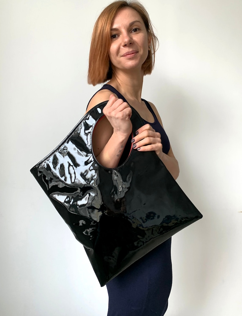 Black patent leather bag is finished with an outside vertical zipper pocket and a strap. The bag is fully lined and has two inner pockets. The bag can be carried on the shoulder or crossbody. Height 15 Width 13.5 inches.