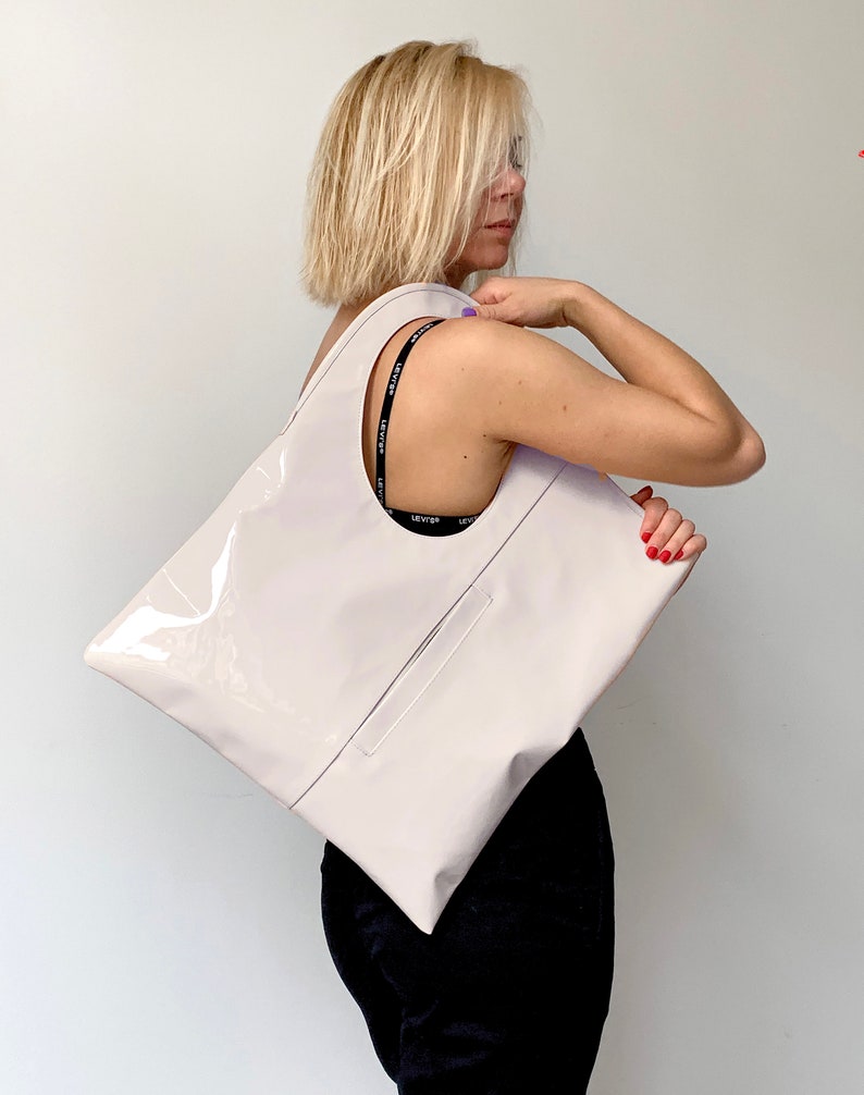 White patent leather shoulder bag is finished with an outside vertical zipper pocket and an adjustable strap. The bag is fully lined and has two inner pockets. The bag can be carried on the shoulder or crossbody. Height 19 Width 17 inches.