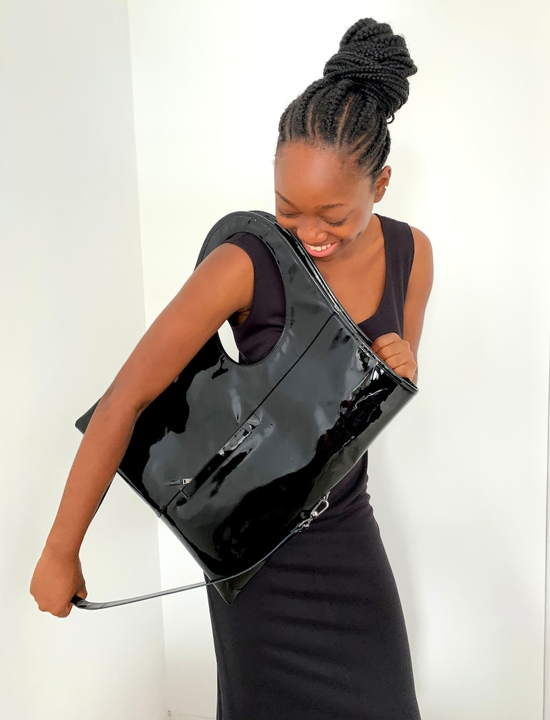 Black patent leather shoulder bag is finished with an outside vertical zipper pocket and an adjustable strap. The bag is fully lined and has two inner pockets. The bag can be carried on the shoulder or crossbody. Height 19 Width 17 inches.