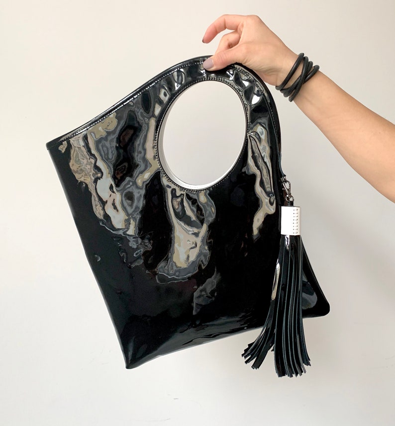 Black patent leather bag is finished with an outside vertical zipper pocket and a strap. The bag is fully lined and has two inner pockets. The bag can be carried on the shoulder or crossbody. Height 15 Width 13.5 inches.