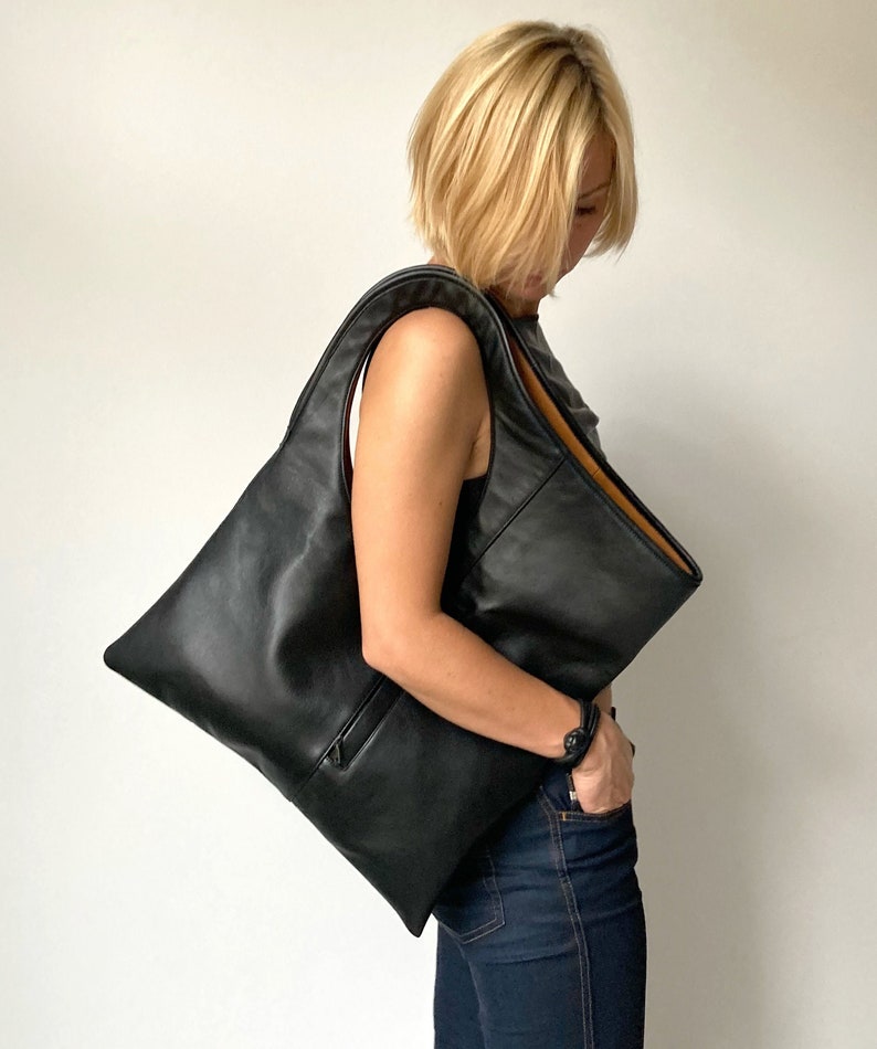 Black leather shoulder bag with tan interior collar. It has a rectangular shape with an oval armhole holder. The bag has an outside vertical zipped pocket. The bag can be carried in hand as a tote, over the shoulder or as a fold over clutch.