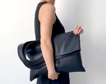 May include: A black leather tote bag with a fold-over flap and a single strap. The bag is being held by a person wearing a black sleeveless dress.