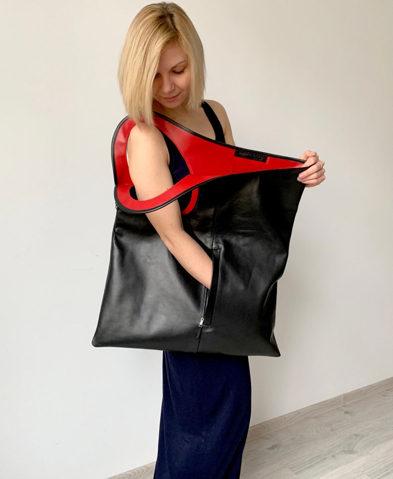 Black leather bag is finished with an outside vertical zipper pocket and a strap. The bag is fully lined and has two inner pockets. The bag can be carried on the shoulder or crossbody. Height 22 Width 20 inches.