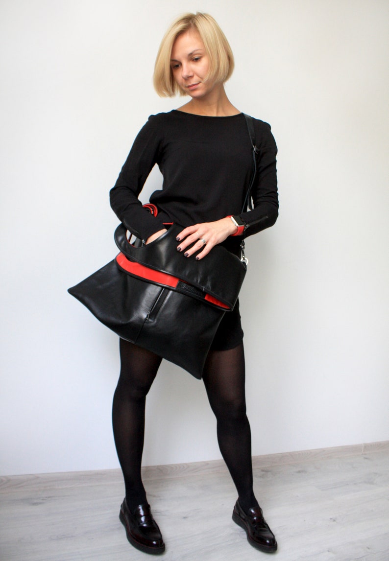 Black leather shoulder bag with red interior collar is finished with an outside vertical zipper pocket and a strap. The bag is fully lined and has two inner pockets. The bag can be carried on the shoulder or as a clutch. Height 19 Width 17 inches.