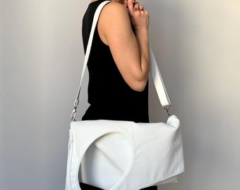 May include: White rectangular shoulder bag with a large circular cutout. The bag has a long, adjustable strap and silver-tone hardware. The bag is made of a smooth material. The person in the image is wearing a black dress.