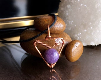 May include: A copper wire wrapped pendant featuring a purple amethyst stone and a clear quartz crystal. The pendant is hanging from a black cord and is resting on brown stones.