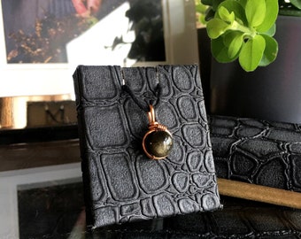 May include: A small, round, dark green gemstone pendant with a copper wire wrap. The pendant is on a black cord and is displayed in a black box with a crocodile skin pattern.