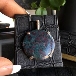 May include: A green and red gemstone pendant with silver wire wrapping. The pendant is displayed on a black display stand with a crocodile pattern.