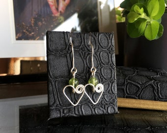 May include: A pair of silver heart-shaped earrings with green glass beads. The earrings are displayed on a black crocodile-embossed surface.