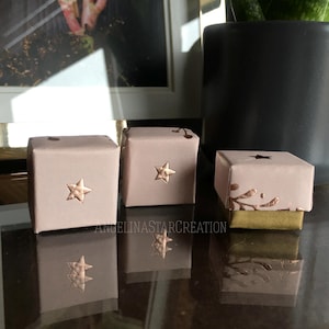 Peut inclure: Trois petites boîtes cadeaux carrées alignées, chacune enveloppée dans du papier rose clair. Une étoile en bronze est fixée sur le devant de chaque boîte. Les boîtes sont sur une surface réfléchissante. Le texte "ANGELINASTARCREATION" est visible.
